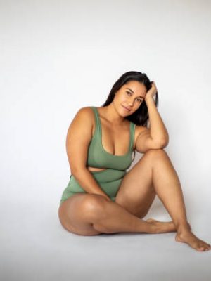 Portrait of a beautiful oversize woman in lingerie sitting on white background. Plus size woman in underwear looking at camera with hand on head.
